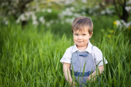 Cheerful little boy with blue eyes in overalls standing in green meadow