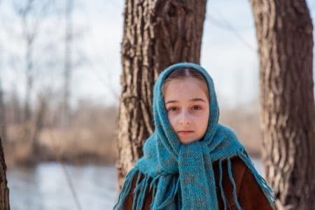 Cute girl wearing sheepskin coat stands near tree by the river