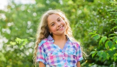 Cheerful pretty young girl with long curly hair smiling to the camera in green meadow on sunny day