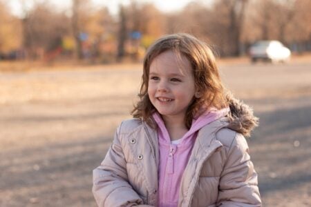 Beautiful red-haired little girl in jacket with a fluffy hood smiles on the background of autumn street