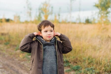 Adorable young boy wearing jacket standing on rough road with grassy field