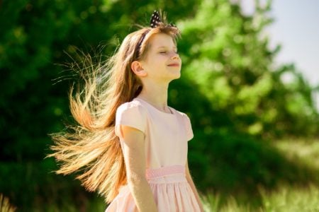 Adorable girl in elegant dress smiling while closed eyes in summer field