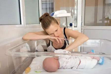 Mother sitting beside her newborn baby sleeping in hospital cradle looking concerned