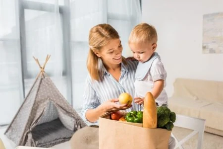 Smiling mother showing the groceries to her baby while carrying him