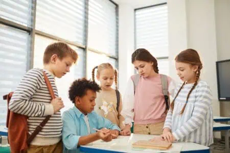 Young boy sitting with group of students standing around him helping with task at school