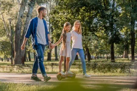 Smiling parents walking in park with their cute little girl riding hoveboard